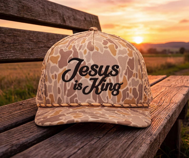 Puede incluir: Una gorra de b&eacute;isbol de camuflaje con un patr&oacute;n en tonos beige, marr&oacute;n y crema, que presenta un cord&oacute;n trenzado en la base de la corona. El panel frontal muestra el texto bordado en negro Jesus is King en una fuente cursiva estructurada.