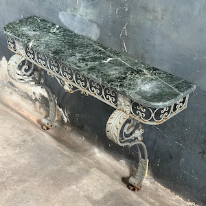 May include: A long, rectangular console table with a green marble top and ornate, scrolled metal legs. The metal frame is painted in a light gray color with gold accents. The table is against a dark, textured wall.