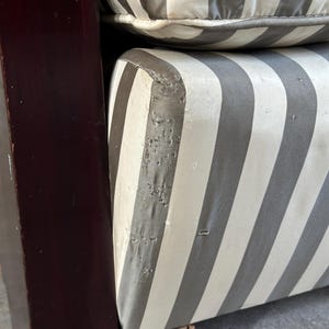 May include: Close-up of a section of a striped upholstered chair. The cushions feature wide vertical stripes in gray and off-white. The wooden frame is a dark, reddish-brown color. The upholstery shows signs of wear and tear.