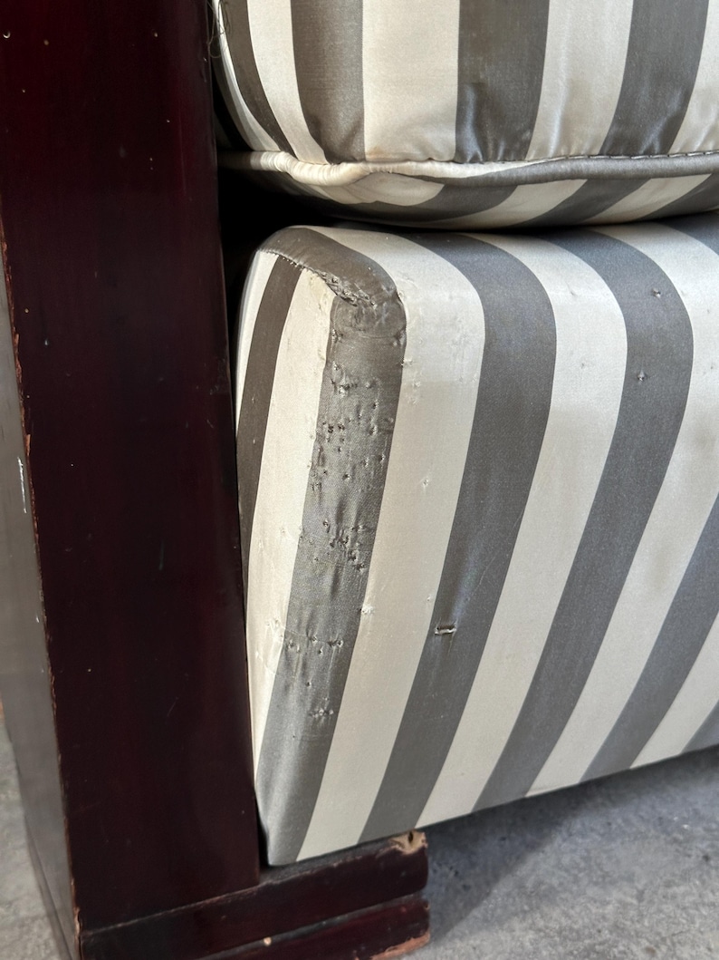 May include: Close-up of a section of a striped upholstered chair. The cushions feature wide vertical stripes in gray and off-white. The wooden frame is a dark, reddish-brown color. The upholstery shows signs of wear and tear.