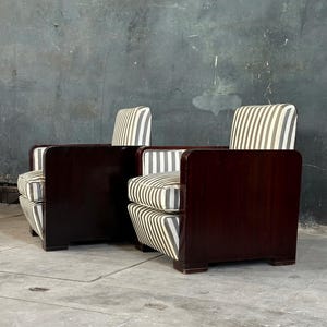 May include: Two Art Deco style armchairs. The chairs have dark wood frames and are upholstered with striped fabric in shades of white, gray, and black. The chairs have a low profile with a rectangular shape and a curved back.