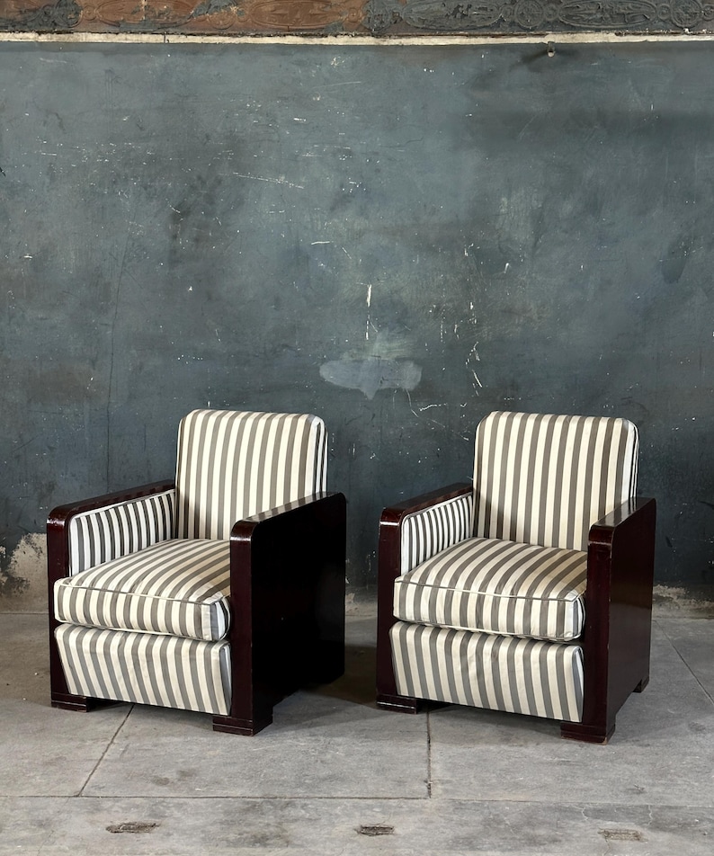 May include: Two matching armchairs with dark wood frames and striped cushions. The cushions feature vertical gray and white stripes. The armrests and frames are a dark, rich wood color. The chairs are set against a dark gray wall.