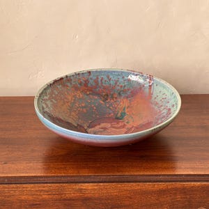 May include: A handmade ceramic bowl with a wide, shallow design. The bowl features a speckled glaze in shades of teal, red, and brown. The rim has a light blue hue, and the interior has a mottled pattern. The bowl is likely used for serving or display.