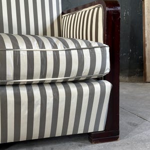 May include: Close-up of a vintage sofa with a striped pattern in gray and white. The cushions and backrest feature the same vertical stripe design. The wooden frame is a dark, rich brown. The sofa has a classic, art deco style.