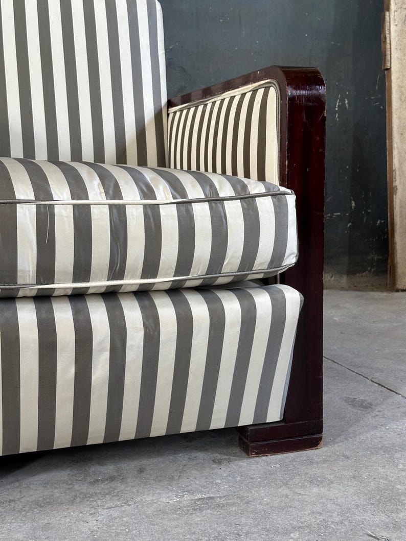 May include: Close-up of a vintage sofa with a striped pattern in gray and white. The cushions and backrest feature the same vertical stripe design. The wooden frame is a dark, rich brown. The sofa has a classic, art deco style.