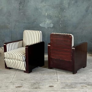May include: Two vintage armchairs. The chairs have dark wood frames with a rectangular design. The seats and backs are upholstered in a striped fabric of gray and white. The chairs are placed on a concrete floor against a dark gray wall.