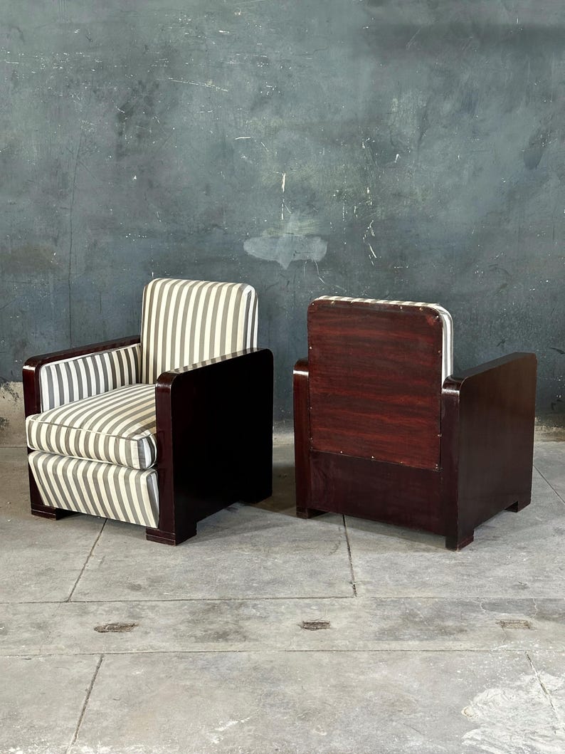 May include: Two vintage armchairs. The chairs have dark wood frames with a rectangular design. The seats and backs are upholstered in a striped fabric of gray and white. The chairs are placed on a concrete floor against a dark gray wall.