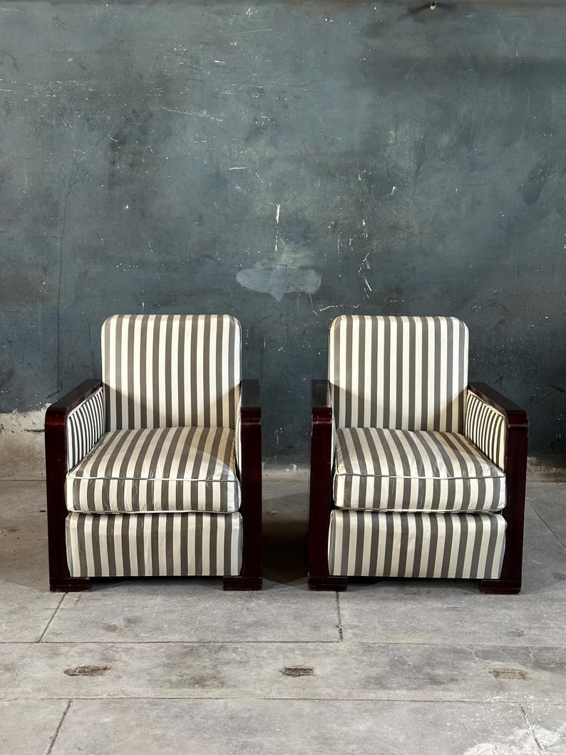 May include: Two matching armchairs with a striped pattern in shades of gray and white. The chairs have dark wood frames and armrests. The background is a textured, dark gray wall.