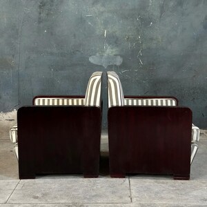May include: Two matching art deco armchairs. The chairs have dark wood frames with a rectangular back and armrests. The seat backs are upholstered in a striped pattern of white and gray. The chairs are placed side by side.