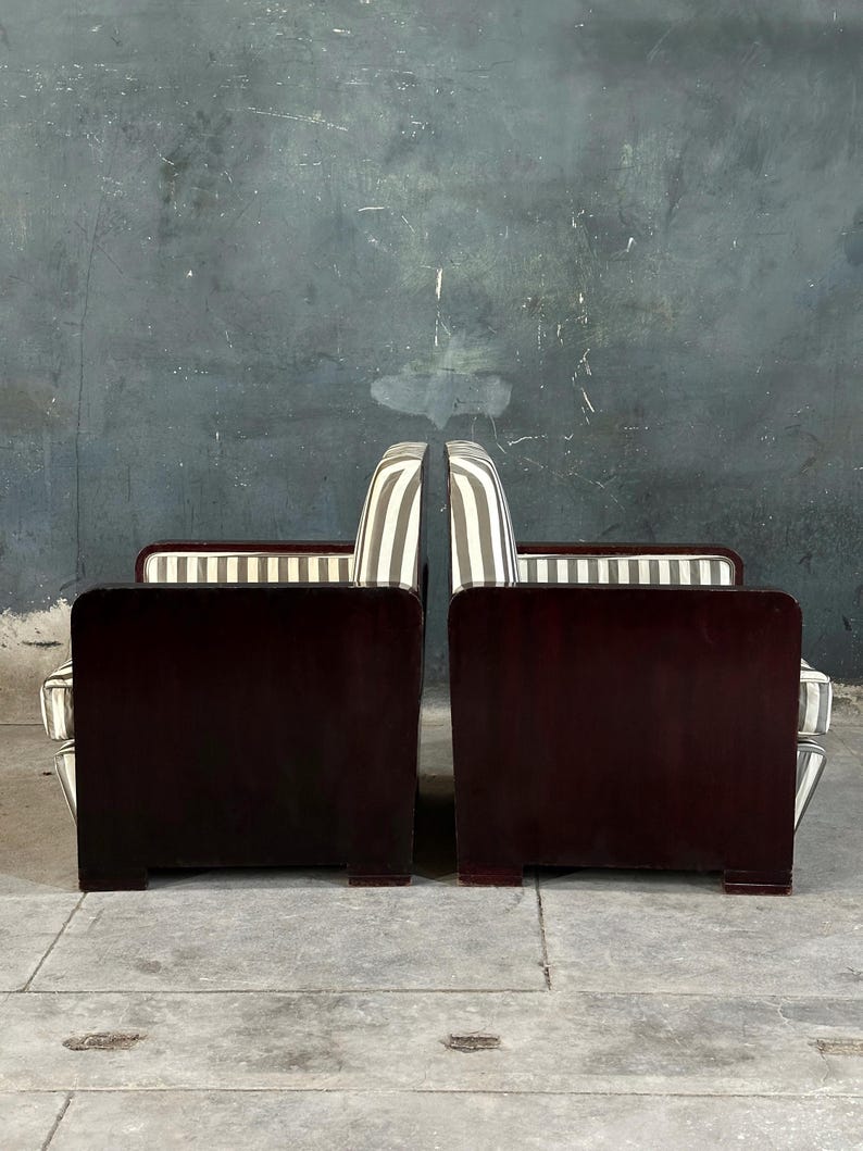 May include: Two matching art deco armchairs. The chairs have dark wood frames with a rectangular back and armrests. The seat backs are upholstered in a striped pattern of white and gray. The chairs are placed side by side.