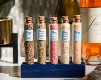 May include: Five small glass bottles with cork tops filled with different colored salts. The bottles are labeled with the text "Boca Sea Salt Co." and the type of salt, including "Smoked Pecan", "Smoked Rosemary Garlic", "Pinot Noir", "Sauv Blanc Thyme & Lemon", and "Rose". The bottles are arranged in a row on a blue wooden stand.