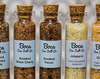 May include: Five small glass bottles with cork tops filled with different flavored salts. The labels read "Boca Sea Salt Co." and include flavors like Smoked Rosemary Garlic, Smoked Black Cherry, Smoked Pecan, Jalapeno, and Diablo.