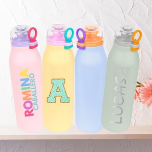 Puede incluir: Cuatro botellas de agua coloridas con textos y diseños personalizados. Las botellas son rosa, amarilla, azul y verde, cada una con un color de tapa diferente y un asa de bucle. Las botellas tienen los nombres "ROMINA CABALLERO" y "LUCAS", y la letra "A".