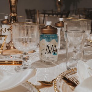 Puede incluir: Una mesa puesta con un n&uacute;mero de mesa de acr&iacute;lico dorado y transparente que dice "Mesa 7". Hay copas de agua de vidrio transparente y una botella de vidrio transparente con tapa dorada y una etiqueta azul y blanca que dice "MAMA".