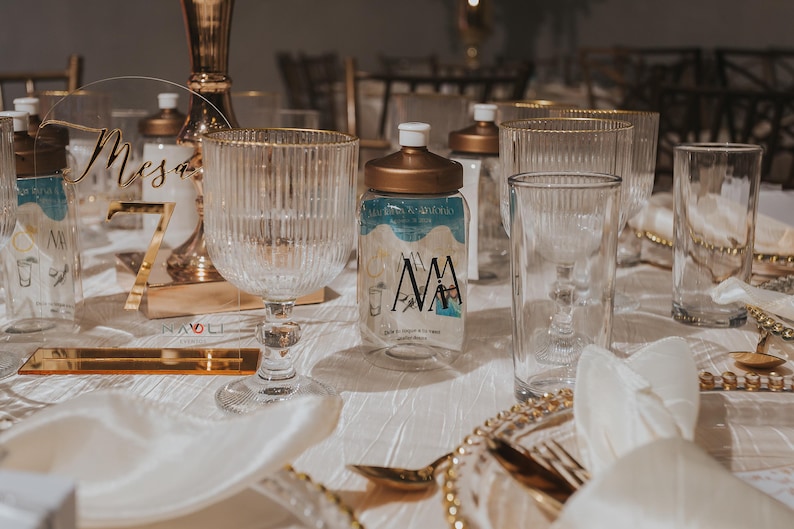 Puede incluir: Una mesa puesta con un n&uacute;mero de mesa de acr&iacute;lico dorado y transparente que dice "Mesa 7". Hay copas de agua de vidrio transparente y una botella de vidrio transparente con tapa dorada y una etiqueta azul y blanca que dice "MAMA".