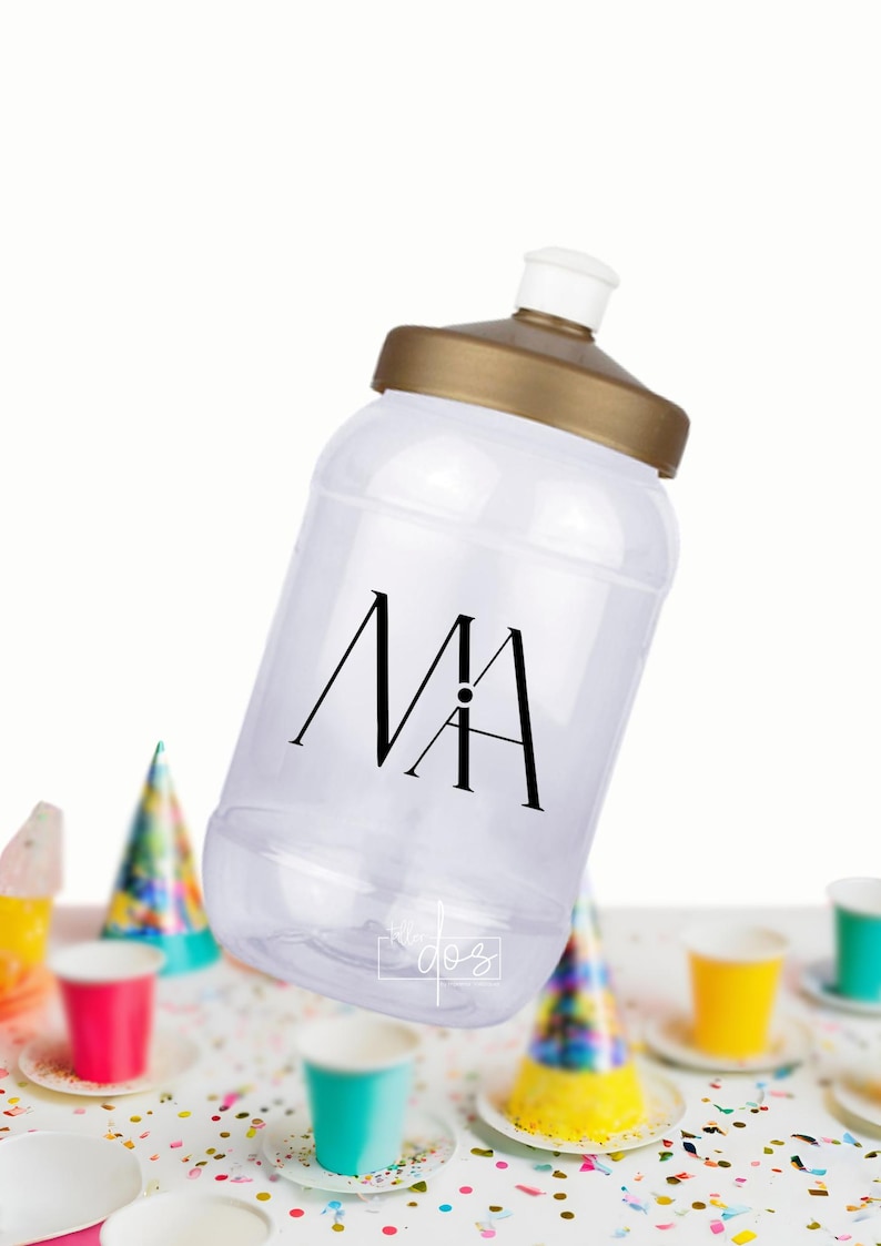 Puede incluir: Una botella de agua de pl&aacute;stico transparente con tapa dorada y un monograma "MA" negro. La botella est&aacute; sobre una mesa con confeti de colores y art&iacute;culos de fiesta.