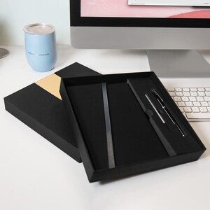Puede incluir: Caja de regalo negra abierta, que contiene un bolígrafo y una regla de metal plateado. Un vaso azul claro con tapa plateada está a la izquierda. El bolígrafo es negro con detalles plateados.