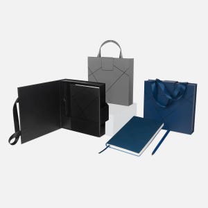 Puede incluir: Un surtido de artículos de papelería, que incluyen un folio negro, una bolsa de regalo gris y una bolsa de regalo azul marino, todos con diseños de líneas geométricas. También hay un cuaderno y un bolígrafo azul marino, lo que sugiere un tema profesional o corporativo.