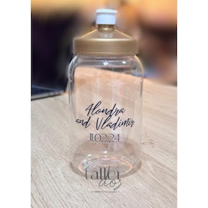 Puede incluir: Botella de agua de pl&aacute;stico transparente con tapa dorada y una etiqueta negra que dice "Alondra y Vladimir 11.02.24".