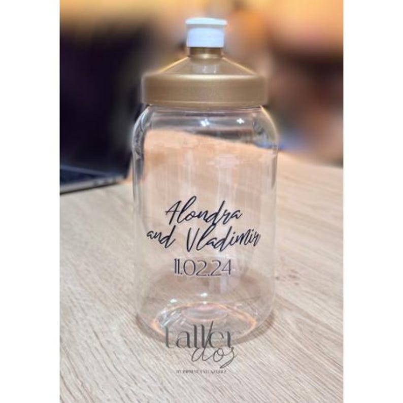 Puede incluir: Botella de agua de pl&aacute;stico transparente con tapa dorada y una etiqueta negra que dice "Alondra y Vladimir 11.02.24".