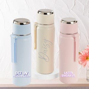 Puede incluir: Tres botellas de agua aisladas en colores pastel: azul claro, crema y rosa. Cada botella tiene una tapa plateada y un asa del mismo color. La botella crema tiene el nombre "Daisy" en cursiva. La botella azul claro dice "DTF UV PERSONALIZADO" y la botella rosa dice "GRABADO LÁSER".