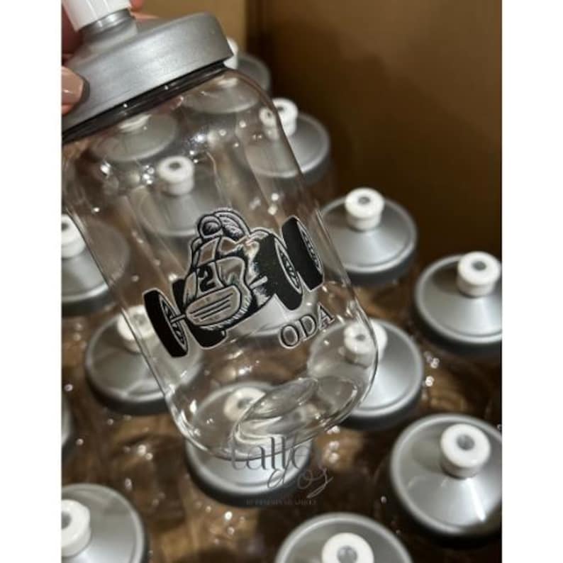 Puede incluir: Una botella de agua de pl&aacute;stico transparente con tapa plateada y un gr&aacute;fico en blanco y negro de un coche de carreras con la palabra "ODA" impresa en el lateral.