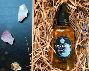 Puede incluir: Una botella de vidrio transparente con una etiqueta marrón que dice "Lullaby Relaxing Body Oil" en una caja de cartón marrón llena de papel triturado. La botella está llena de un líquido dorado.