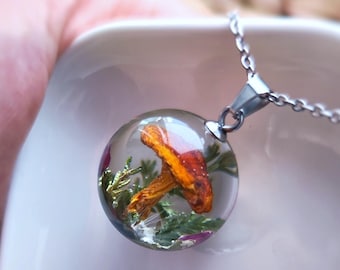 May include: A clear, round pendant necklace featuring a preserved orange mushroom and greenery encased in resin. The pendant hangs from a silver-colored chain. The necklace is held in a white square dish.