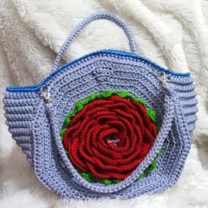 Pode incluir: Uma bolsa de ombro de croch&ecirc; com uma borda azul e uma grande rosa vermelha na frente. A bolsa tem uma al&ccedil;a &uacute;nica e &eacute; feita de um fio macio e texturizado.