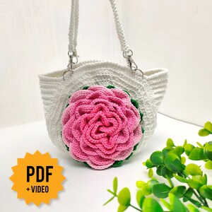 Pode incluir: Sacola tote branca de croch&ecirc; com uma rosa de croch&ecirc; rosa e verde na frente. A bolsa tem duas al&ccedil;as brancas com fechos prateados. O texto "PDF + VIDEO" est&aacute; em uma estrela amarela.
