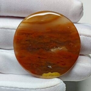 May include: A polished, round gemstone with a warm color palette. The stone features bands of orange, brown, and white, creating a layered effect. The gemstone is held by a white gloved hand.