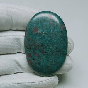 Può includere: Una pietra preziosa verde liscia di forma ovale con inclusiones rosse e bianche. La pietra è lucidata e ha un aspetto screziato.