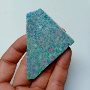 Könnte beinhalten: Ein dreieckiges Stück grüner und blauer Edelstein mit rosa Einschlüssen. Der Edelstein ist roh und hat eine natürliche, unpolierte Oberfläche.