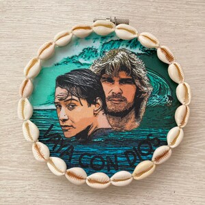 Puede incluir: Un tapiz de pared redondo con un borde de conchas marinas. El centro presenta una imagen colorida de dos hombres con el texto "La Concha" impreso debajo.