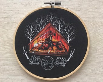 May include: A round, black fabric embroidery hoop featuring a detailed illustration. The image depicts a red cabin with a figure inside, surrounded by pumpkins and leafless white trees. A white sigil is at the bottom.