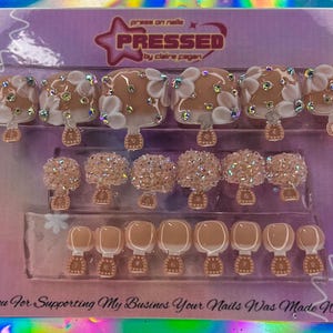 May include: A set of press-on nails in a clear plastic package. The nails feature a light brown base with white floral designs, rhinestones, and a decorative base. The packaging includes the text "PRESSED by claire pagan" and "Thank You For Supporting My Business Your Nails Was Made With Love."