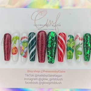 May include: A set of ten long, rectangular press-on nails with a Christmas theme. Designs include red glitter, green glitter, candy cane stripes, and Grinch character art. The nails are displayed on a white card with text that reads "GLOWGIRLS BLUSH".