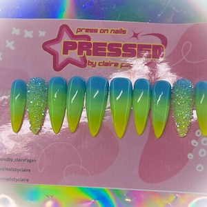 May include: A set of ten long, stiletto-shaped press-on nails with a gradient design. The nails are yellow at the base, transitioning to green, and then blue at the tips. One nail features a cluster of clear rhinestones.