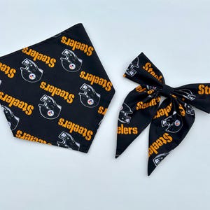 May include: A black triangular bandana and a matching hair bow, both featuring a repeating pattern of yellow Steelers text and black football helmet graphics with the team logo. The fabric is black with a consistent print of the team branding.