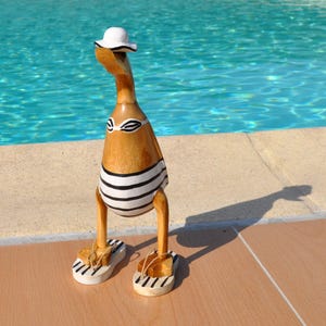 Peut inclure: Figurine de canard en bois portant un chapeau de soleil blanc, un haut de bikini à rayures noires et blanches et un bas assorti. Le canard porte également des sandales à rayures blanches et noires. La figurine est près d'une piscine.