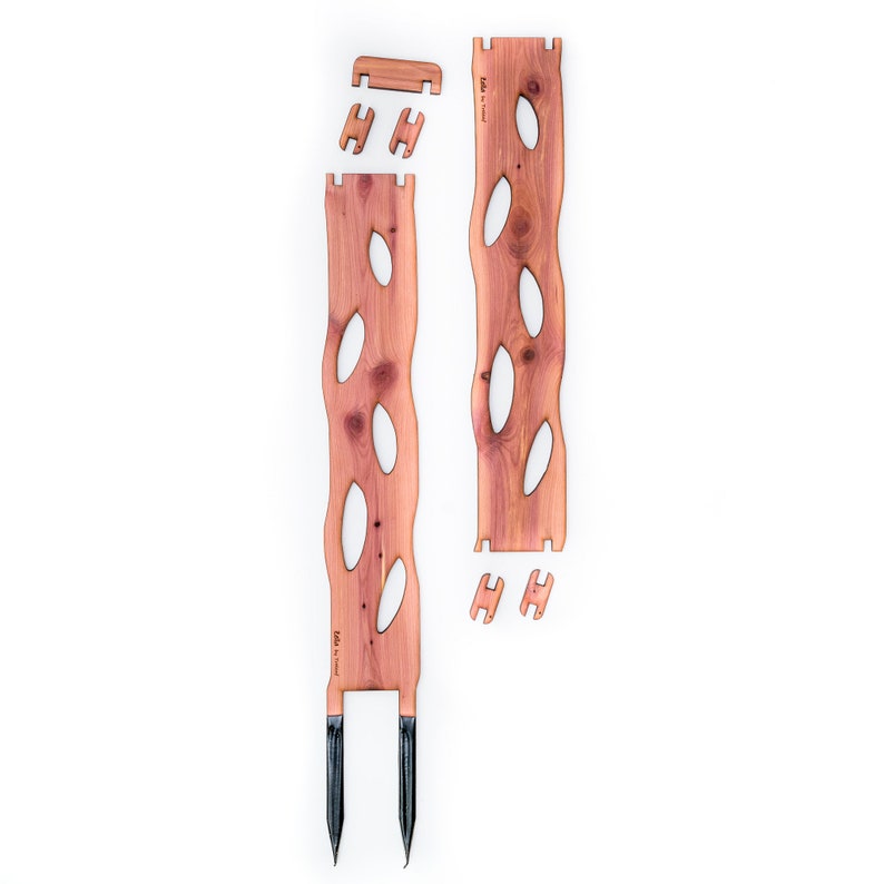 May include: A disassembled wooden garden trellis kit. The kit includes two long, reddish-brown cedar planks with oval cutouts, several smaller pieces, and two black metal stakes. The pieces are arranged on a white background.
