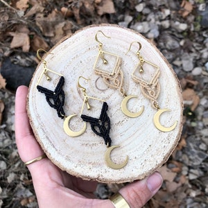 Op de afbeelding: Vier paar handgemaakte oorbellen met een hemels thema. Elke oorbel heeft een goudkleurig vierkant frame, macramé details en een halve maan bedel. Twee paar zijn zwart en twee zijn beige, allemaal met goudkleurige oorhaken.