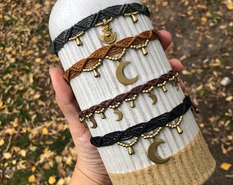 May include: Four macrame chokers in gray, brown, and black, each adorned with gold-colored beads and charms. The charms include crescent moons and a pendant. The chokers are displayed on a white and tan ceramic vase.