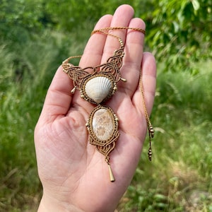 Könnte beinhalten: Eine Makramee-Halskette mit einer weißen Muschel und einem ovalen, versteinerten Korallen-Cabochon in brauner Kordel. Das Design umfasst komplizierte Knoten, kleine Metallperlen und einen hängenden, tropfenförmigen Metallanhänger.