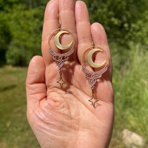 Könnte beinhalten: Ein Paar Ohrhänger mit einem goldfarbenen Halbmond in einem kreisförmigen Rahmen, akzentuiert durch ein geknotetes mauvefarbenes Makramee-Band und einen kleinen vierzackigen Sternanhänger, der an der Unterseite jedes Ohrrings hängt.