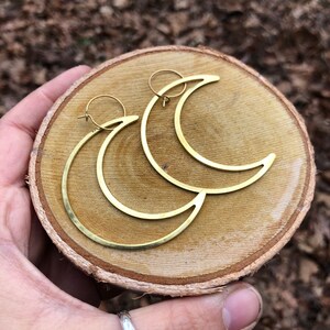 Könnte beinhalten: Ein Paar goldfarbene Halbmond-Ohrringe. Die Ohrringe werden auf einer runden, hellbraunen Holzoberfläche präsentiert. Die Ohrringe haben ein schlichtes, minimalistisches Design mit einem Creolenverschluss.