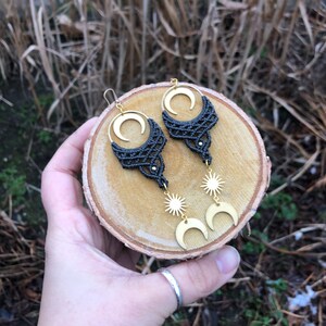 Op de afbeelding: Een paar handgemaakte macramé oorbellen met een maan- en zonontwerp. De oorbellen hebben een donkergrijs geweven patroon met goudkleurige metalen accenten. De oorbellen worden op een houten oppervlak gepresenteerd.