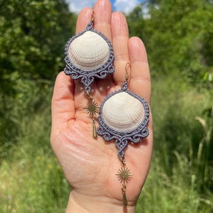 May include: A pair of earrings featuring white seashell centres framed by intricate grey macrame cord. Each earring includes a gold-toned metal sunburst charm and a small teardrop-shaped pendant hanging from the bottom of the shell structure.