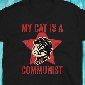 以下が含まれることがあります： 赤い星と赤い帽子をかぶった猫が描かれた黒いTシャツ。Tシャツには「My cat is a communist」という文字がプリントされています。