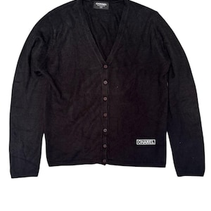 May include: Black cardigan sweater with a V-neck and button-down closure. The sweater has long sleeves and a small white logo patch that says "CHANEL". The brand name is also on the label.